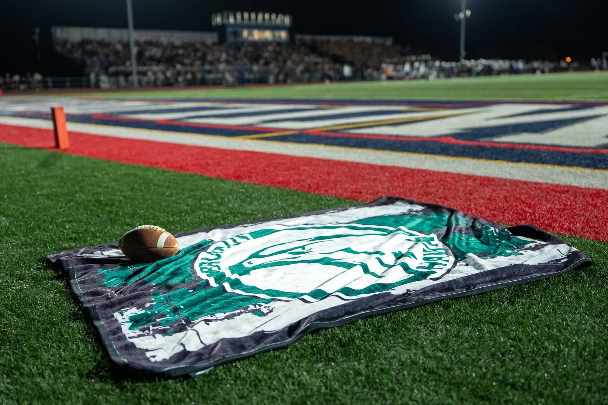 A custom mascot blanket and a football laid in the end zone under the lights, packed home stands filling the background